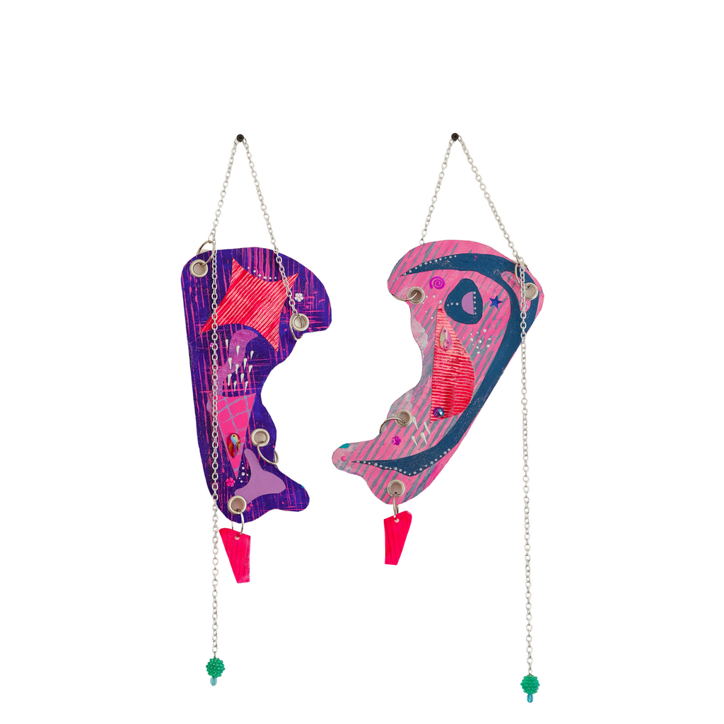 The artwork 'Rise Above' by Sunny Chapman features two abstract hanging shapes with vibrant purple and pink tones, embellished with metallic chains and decorative elements. Each piece showcases intricate mixed media details including glitter, sequins, and jewels, suspended by silver chains with teal bead accents and bright pink dangling elements.