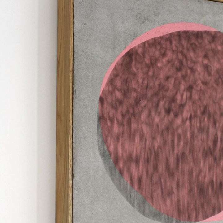 The artwork 'Roller Skating Example' by Evan Ishmael features a large textured rose-pink circle on a muted gray concrete background. The piece combines soot and spray-applied acrylic on panel, creating a dynamic contrast between the rough, organic texture of the circle and the smooth industrial surface. The wooden frame complements the contemporary minimalist composition.