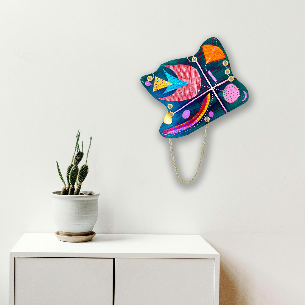 The artwork 'Quartet' by Sunny Chapman features a vibrant butterfly wall hanging with bold geometric patterns. Deep teal wings adorned with pink, orange, and yellow shapes create a playful abstract design. Gold accents and curved lines add dynamic movement. The piece combines acrylic paint on paper in a 16x12 inch format. Art by Sunny Chapman.