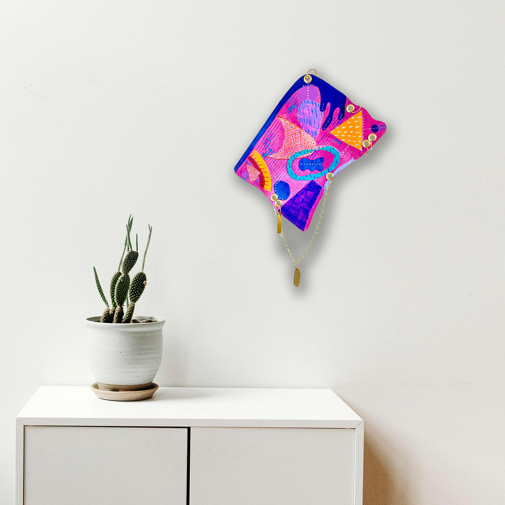 The artwork 'Seaside' by Sunny Chapman features a vibrant abstract composition with flowing organic shapes in neon pink, electric blue, and golden yellow. The piece displays a dynamic, psychedelic-inspired pattern mounted on a wall above a potted cactus. Acrylic on paper, the artwork creates a striking contrast against the white background. By Sunny Chapman.