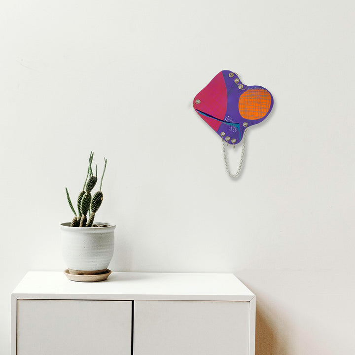 The artwork 'Satellite' by Sunny Chapman features an abstract geometric composition in vibrant magenta, purple, and orange hues on paper. The design incorporates curved shapes connected by a delicate silver chain, creating a modern, playful wall piece. The 12x12 inch acrylic painting contrasts boldly against a white background, by Sunny Chapman.