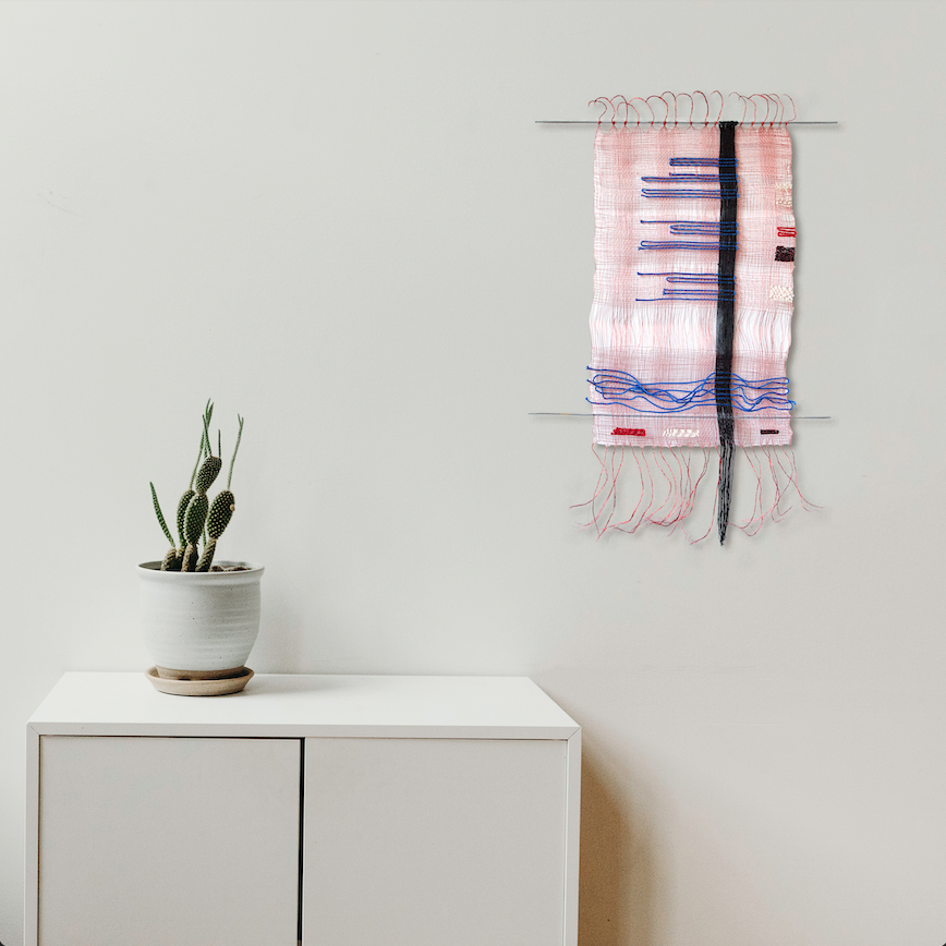 The artwork 'Svo-bo-dy! #1, Russian Morse Code' by Kathie Halfin features a delicate wall hanging with pink cotton fabric adorned with blue and red horizontal stripes in morse code pattern. A vertical black rope bisects the piece, while copper wire and pink flux yarn create textural fringe elements. Displayed alongside a minimalist white cabinet with a small cactus plant.