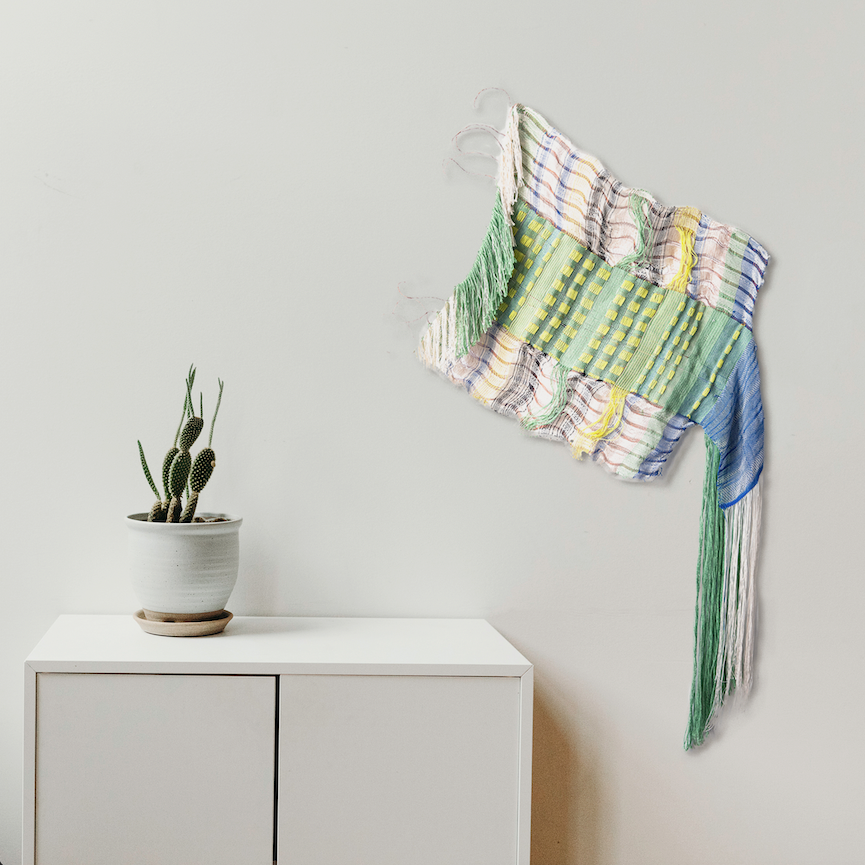 Loose, English Morse Code by Kathie Halfin: Abstract wall textile featuring interwoven cotton and flux yarn with copper wire elements. Displays a delicate interplay of mint green, blue, and white hues. Flowing fringe details create dynamic movement while geometric patterns suggest morse code elements. Mounted beside a minimalist white cabinet with a small cactus plant.