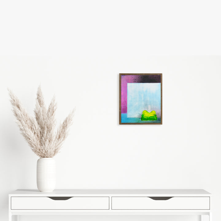 The artwork 'Snow home experience' by Evan Peltzman features abstract geometric shapes in vibrant colors on a wood panel. A serene light blue background contrasts with a bold magenta edge, while a bright lime green form rests at the bottom. The mixed media piece combines ink and plaster textures, creating depth within its framed composition.