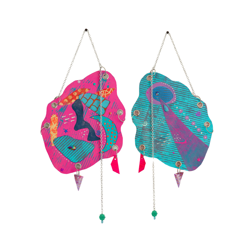 The artwork 'Space Oddity' features two hanging abstract panels in vibrant pink and turquoise, adorned with celestial and aquatic motifs, metallic chains, beads, and decorative elements. Mixed media piece combining acrylic paint, glitter, sequins, and handmade charms creates a whimsical cosmic-meets-ocean aesthetic. By Sunny Chapman