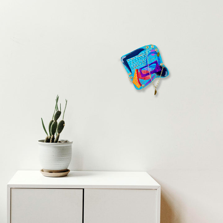 The artwork 'Spate' by Sunny Chapman features vibrant abstract shapes in electric blue, magenta, and orange tones creating a dynamic floating composition against a white wall. The 12x12 inch acrylic on paper piece displays fluid, energetic brushstrokes with a contemporary aesthetic, positioned above a minimalist white cabinet with a small potted cactus.