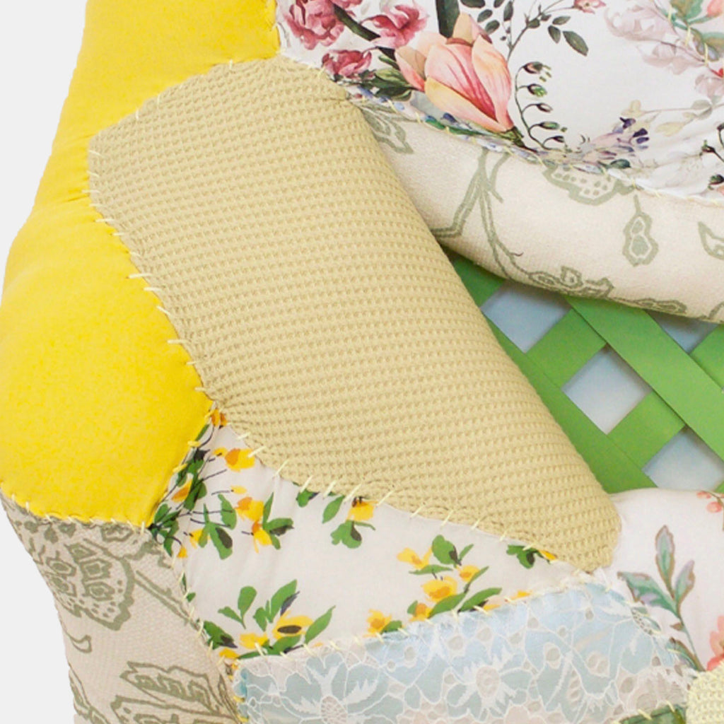 The artwork 'Spring Fever' by Hanna Washburn features a textural assemblage of recycled fabrics featuring floral patterns in yellow, pink, and blue hues, arranged against a green wooden lattice. Delicate quilting and stitching details create dimensional interest, while vintage textiles showcase delicate flower prints and geometric textures, embodying a fresh springtime aesthetic.