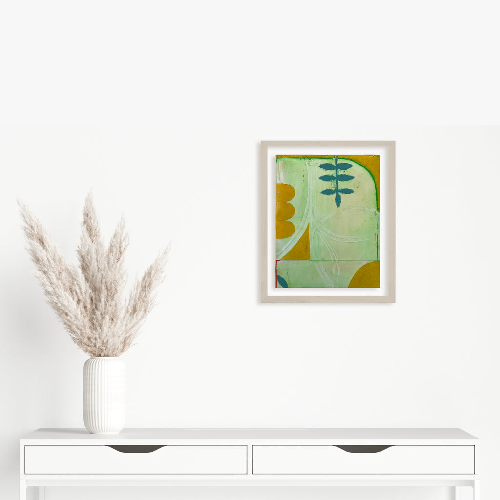 Spring Fling 5 by Janice La Motta features abstract organic shapes in mint green and golden yellow, with a delicate botanical motif resembling a fern branch. Created with oil pastel on Yupo paper, the composition balances soft curves with geometric elements in a minimalist style, framed in white. Original artwork by Janice La Motta.