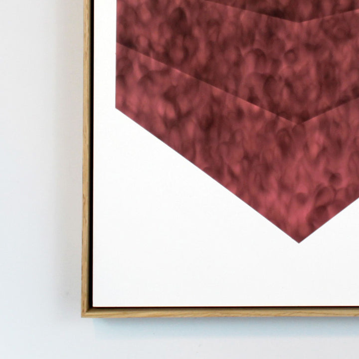 The artwork 'Sunlight In The House' by Evan Ishmael features a striking geometric shape in deep burgundy tones created with soot and spray-applied acrylic. The angular form appears as an inverted pyramid or chevron against a stark white background, with a textured, mottled surface that creates depth and visual interest. The piece is presented in a minimalist wooden frame, showcasing the contrast between the rich color and negative space.