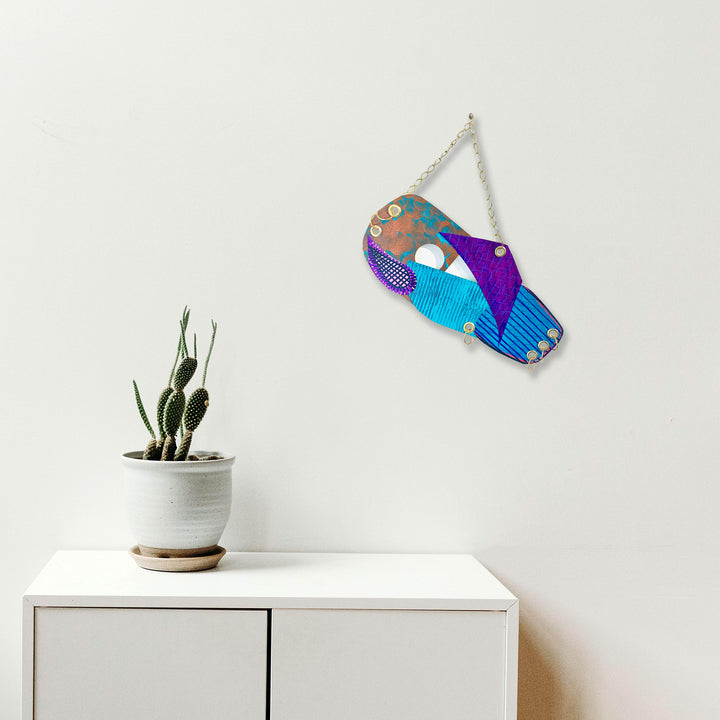 The artwork 'Swinging' by Sunny Chapman features a vibrant abstract fish-shaped wall hanging in turquoise, purple, and copper tones. The decorative piece is crafted in acrylic on paper, displaying a whimsical patchwork pattern with geometric details and metallic accents. Suspended by a silver chain against a white wall, it creates a playful artistic focal point above a potted cactus plant.