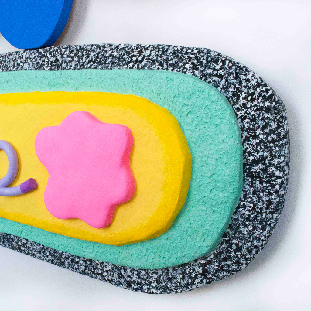 The artwork 'Tasty' by Ryan Patrick Martin features a vibrant abstract composition with layered concentric shapes in blue, turquoise, yellow, and pink. The textured surface combines speckled black and white borders with smooth central elements, creating depth through mixed media including wood and epoxy clay. The whimsical, organic forms suggest playful movement and dimensional exploration.