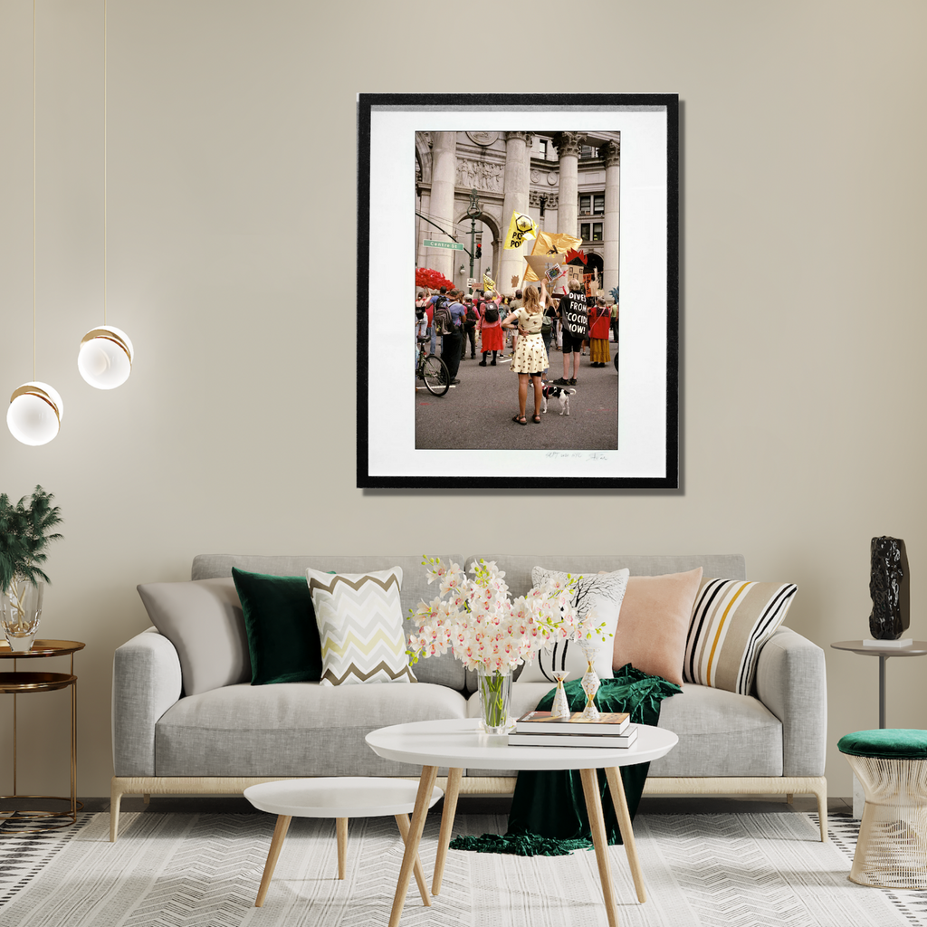 The artwork 'The Argument' by Mari Sarai captures a vibrant street scene with a large yellow balloon against classical architecture backdrop, featuring red umbrellas and a gathering crowd. The vintage-style photograph is presented in a black frame, showing an urban moment in nostalgic sepia-tinted colors. 40x32 inches framed limited edition print by Mari Sarai.