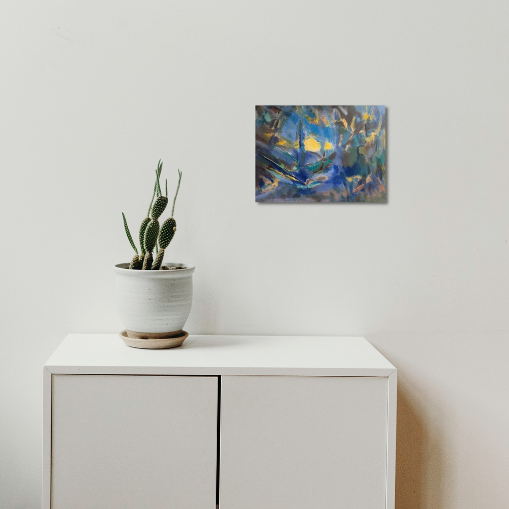 The artwork 'Through the Light' by Beth Barry features ethereal brushstrokes in deep blues and golden yellows, creating an abstract landscape with dynamic movement. The acrylic painting on canvas captures a dreamy, atmospheric quality with fluid forms and luminous effects, displayed above a minimalist white cabinet with a small cactus plant.