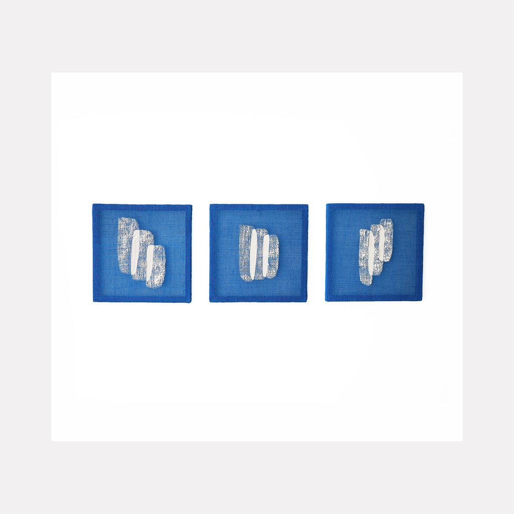 The artwork 'Time Isnt Real I' by Morgan Hale features three square panels in vibrant blue linen, each displaying abstract white plaster formations arranged horizontally. The textural elements appear as geometric columns or bars against the rich blue background, creating a minimalist triptych composition. Handwoven linen and plaster, 9x9 inches. By Morgan Hale.