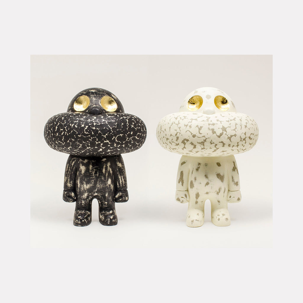 The artwork 'Gatekeeper' by Yusuke Ochiai features two whimsical sculptural figures with mushroom-shaped heads, one in black and one in white cement. Both characters have striking gold leaf eyes and textured, crackle-pattern surfaces. The figures stand side by side, displaying a minimalist yet playful design with rounded forms and simple limbs. Spray-painted cement with gold leaf details.