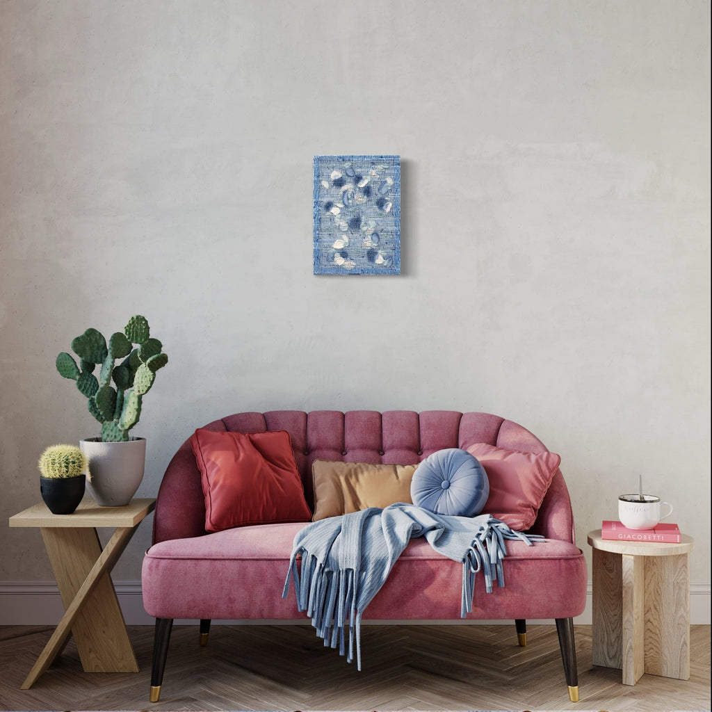 The artwork 'Frost' by Morgan Hale features a textural abstract composition in soft blue and white, crafted from handwoven linen and plaster. The 11x18 inch piece creates a delicate pattern reminiscent of winter frost formations, displayed above a pink tufted sofa with decorative throw pillows and a cactus plant, showcasing its versatility in modern home decor settings.