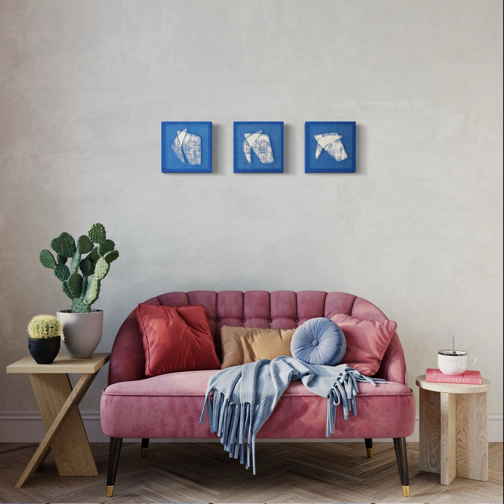 The artwork 'Time Isnt Real III' by Morgan Hale features three square blue panels with abstract white textural forms, displayed above a pink tufted sofa. The 9x9 inch handwoven linen and plaster pieces create a striking geometric composition against a neutral wall, showcasing modern minimalist design through texture and contrast.