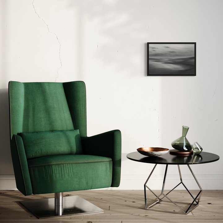 The artwork 'Oceano Dunes ICM' by Neil Shapiro features a minimalist black and white seascape photograph displayed in a black frame, capturing ethereal waves and misty coastal atmosphere using intentional camera movement technique. The framed piece appears on a white wall above a modern interior setting with a emerald green velvet armchair and round side table.