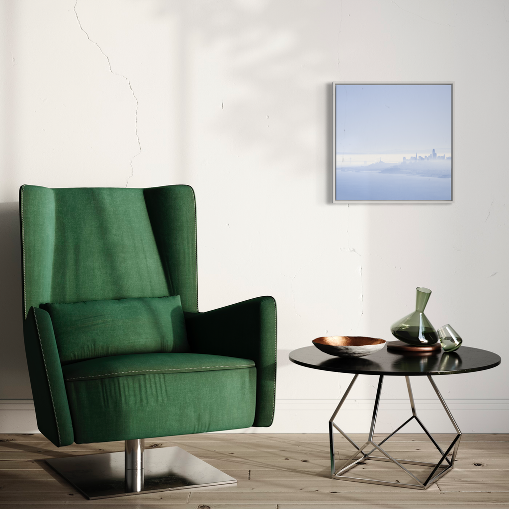 The artwork 'Brumes Matinales' by Fabienne Sowa-Dobkowski captures ethereal morning mist over a minimalist winter landscape, rendered in soft blues and whites. Framed photography showcases serene atmospheric conditions with subtle gradient tones. 20x20 inch archival luster print displayed in modern interior setting with emerald velvet chair. By Fabienne Sowa-Dobkowski.