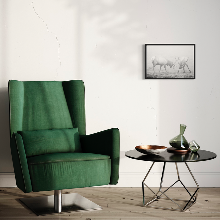 The artwork 'Elks at Play' by Fabienne Sowa-Dobkowski features a black and white framed photograph of elk in a misty setting, displayed on a white wall above a modern side table. The archival luster print showcases the photographer's mastery in capturing wildlife. The 13x19 inch framed piece creates a striking contrast against emerald velvet armchair in a contemporary interior setting.