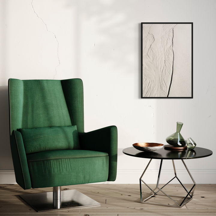 The artwork 'Low Tide' by Fabienne Sowa-Dobkowski showcases a minimalist black-framed photograph displayed in a modern interior setting alongside an emerald green velvet wingback chair and contemporary round side table. The monochromatic print features subtle textural waves and organic lines on archival luster paper, creating a serene coastal aesthetic. - By Fabienne Sowa-Dobkowski