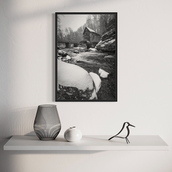 The artwork 'A Winter's Dream' captures a serene black and white photograph of a rustic mill house beside a snow-covered stream. Snow-laden rocks and frozen water create dramatic textures in the foreground, while bare winter trees frame the weathered structure in the background, creating a nostalgic winter landscape. Photography by Dennis Maida.