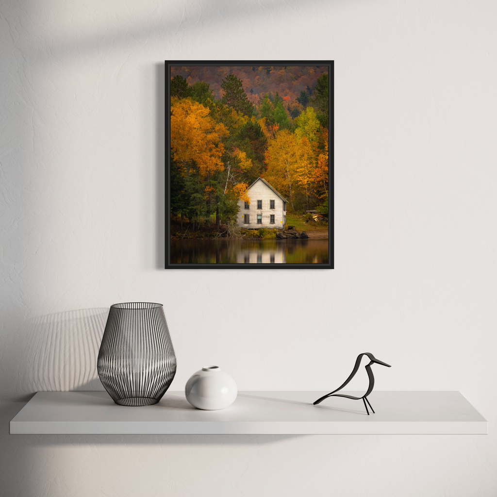 The artwork 'Adirondack House' by Dennis Maida captures a serene white two-story house nestled among vibrant autumn foliage in golden yellows and deep greens, reflected perfectly in a still lake. The photographic composition showcases dramatic fall colors against a moody sky, with the solitary house creating a peaceful focal point in the landscape.