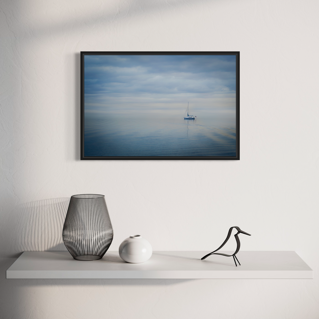 The artwork 'Blue Hour' by Dennis Maida captures a serene seascape photograph featuring a solitary sailboat floating on calm waters. The composition showcases a minimalist scene with muted blue tones merging between sky and sea, creating a dreamy atmospheric effect. The tranquil waters reflect the cloudy sky above, while the distant boat adds a sense of scale and solitude to this peaceful maritime scene.