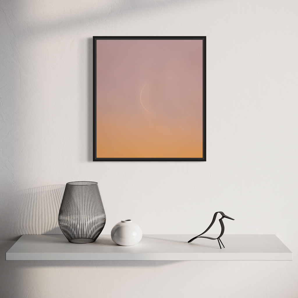The artwork 'C Moon' by Dennis Maida features a minimalist photographic composition with a subtle gradient from soft pink to warm peach tones, displayed in a sleek black frame. Below, a modern vignette showcases decorative objects including a wire-frame vase, white ceramic sphere, and stylized bird figure on a clean white shelf.