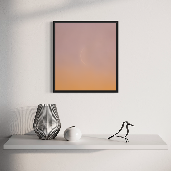 The artwork 'C Moon' by Dennis Maida features a minimalist photographic composition with a subtle gradient from soft pink to warm peach tones, displayed in a sleek black frame. Below, a modern vignette showcases decorative objects including a wire-frame vase, white ceramic sphere, and stylized bird figure on a clean white shelf.