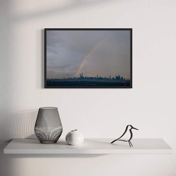 The artwork 'Chasing Rainbows' by Dennis Maida captures a dramatic urban skyline beneath stormy gray clouds, with a vibrant rainbow arching over the city's silhouetted skyscrapers. The atmospheric photograph contrasts the moody darkness of the sky with the hopeful presence of the rainbow, creating a compelling narrative of nature's beauty amid the metropolitan landscape.
