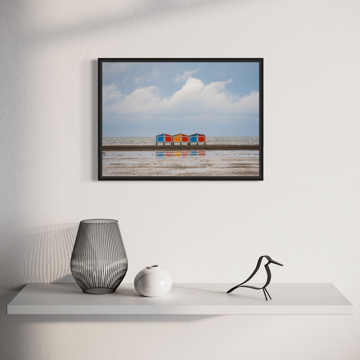 The artwork 'Sand and Sky' by Dennis Maida captures a minimalist coastal scene featuring four vibrant beach chairs - blue, red, and orange - arranged in a row against a concrete seawall. The composition contrasts the chairs' bold colors with a serene backdrop of soft blue sky and fluffy white clouds, creating a striking balance of urban and natural elements.