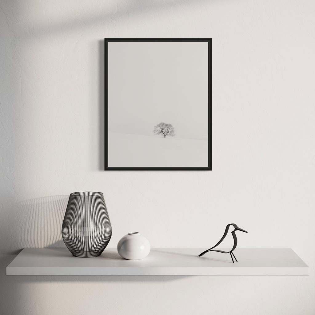 Silent Witness to Winter's Awakening by Dennis Maida: A minimalist black and white photograph featuring a solitary tree in a misty winter landscape, displayed in a black frame above a modern shelf decorated with a wire-frame vase, white ceramic sphere, and stylized bird sculpture. The composition emphasizes negative space and tranquil simplicity.