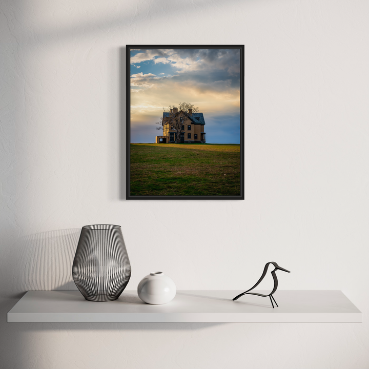 The artwork 'The House Where No One Lives' by Dennis Maida captures a haunting abandoned Victorian house silhouetted against a dramatic sunset sky. The solitary structure stands atop a bare hilltop surrounded by golden fields. Dark, weathered architecture contrasts with luminous clouds in shades of amber and blue, creating an atmospheric fine art photograph of rural solitude.