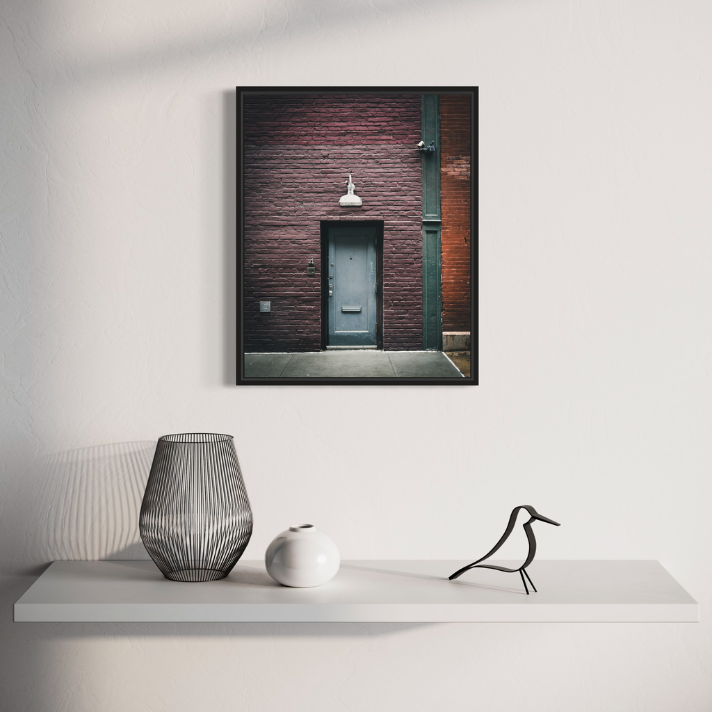 The artwork 'Thresholds Untold' by Dennis Maida captures a minimalist urban scene featuring a teal door set against a textured burgundy brick wall. A vintage-style pendant lamp hangs above the doorway. The composition emphasizes architectural details and geometric lines, creating a moody atmospheric photograph that blends industrial and vintage elements.