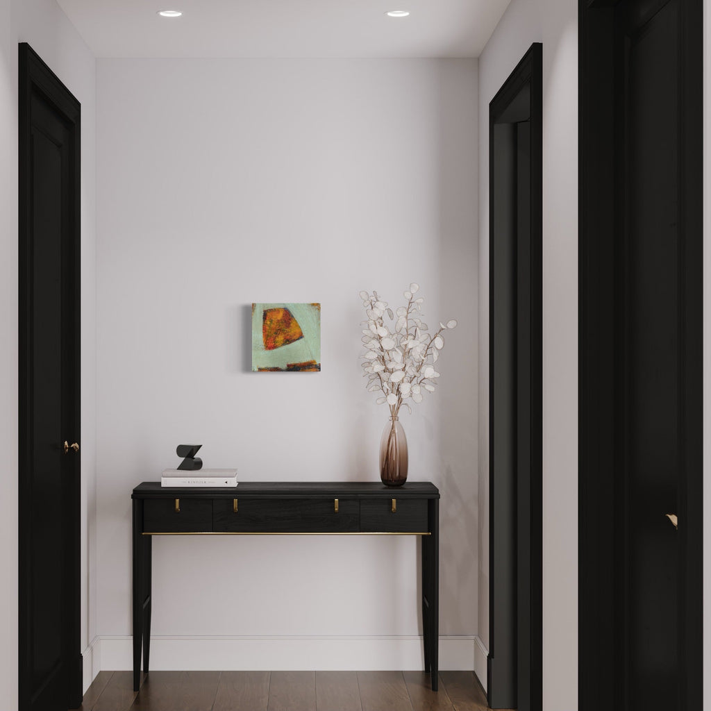 The artwork 'Thinking about differences 3' by Marta Nowicka displayed in a minimalist hallway setting, featuring abstract geometric shapes in orange and mint green tones on a 12x12 inch cradle panel. The acrylic painting creates a striking contrast against the white wall, mounted above a sleek black console table with decorative dried flowers.