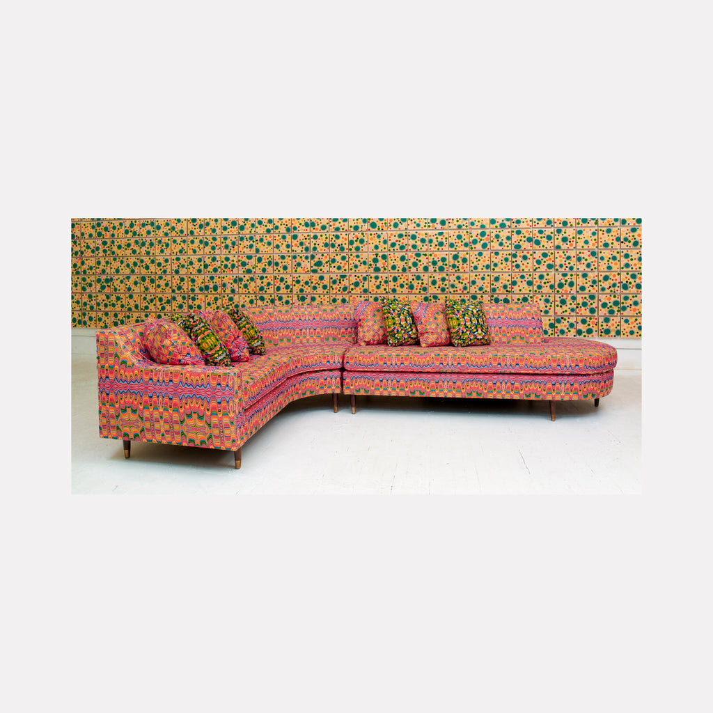 The artwork 'Piece of Cake 887' by Lori Kirkbride features a vibrant L-shaped sectional sofa upholstered in pink and orange patterned fabric with geometric designs. The sofa sits against a retro-style wallpaper with green and gold circular patterns. The piece showcases acrylic polymer and resin on panel, capturing mid-century modern furniture design with contemporary artistic flair.
