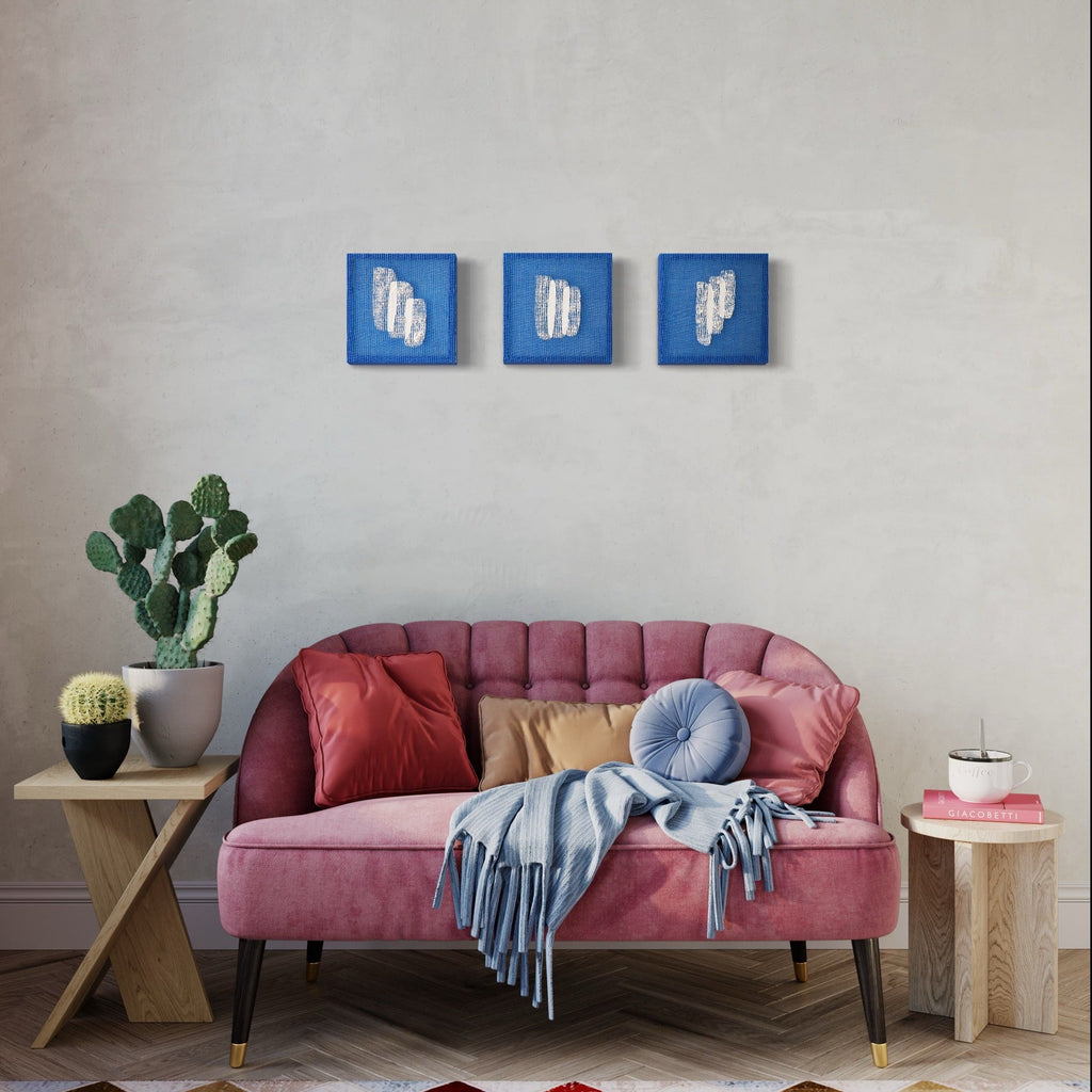 The artwork 'Time Isnt Real I' by Morgan Hale features three square panels with bright cobalt blue backgrounds and abstract white plaster forms. Each handwoven linen panel displays textured, geometric reliefs creating a modern triptych composition. The 9x9 inch artworks showcase a minimalist aesthetic with raised sculptural elements. By Morgan Hale.