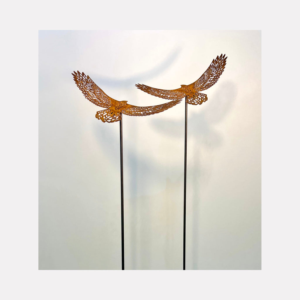 The artwork 'Birds Eye View Tall Earth' by Nicki Adani features two majestic steel birds with intricately detailed wings, rendered in warm copper-toned patina. The birds appear suspended in mid-flight atop tall, slender black steel poles against a soft, ethereal white background, creating a striking 53-inch tall sculptural installation. By Nicki Adani.