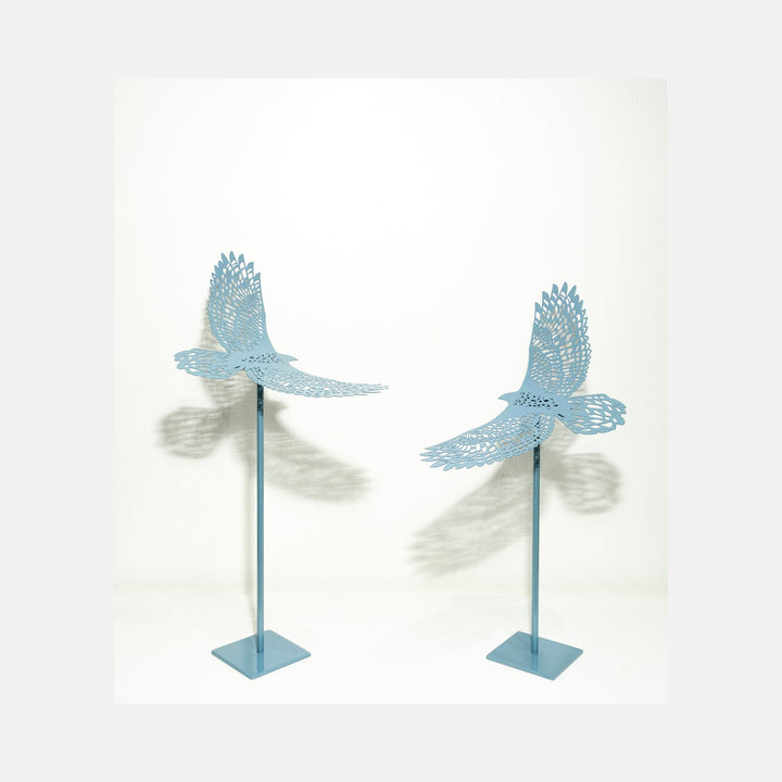 The artwork 'Birds Eye View Sky' by Nicki Adani features two powder-coated steel sculptures of birds in flight, rendered in a soft sky blue finish. Each bird is intricately detailed with delicate wing patterns and mounted on slender vertical stands, casting subtle shadows that enhance their ethereal presence. The contemporary metal art piece displays graceful movement and precision craftsmanship.
