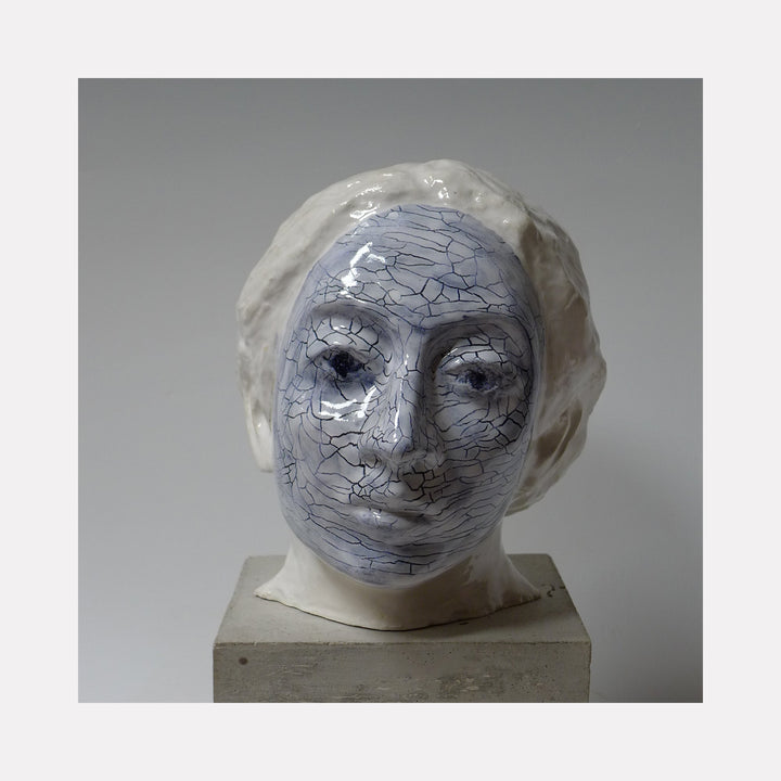 The artwork 'Blue Geraldine' by Mark LaRiviere features a sculptural ceramic head with a distinctive crackled blue-glazed face pattern and smooth white textured hair. The glossy ceramic piece exhibits a striking contrast between the cool blue tones and white surfaces, showcasing intricate surface detailing and artistic craftsmanship. By Mark LaRiviere.