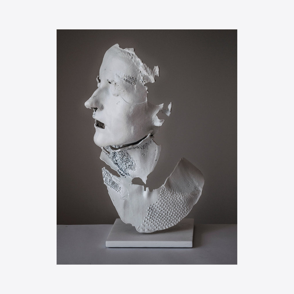 The artwork 'Bust of a Woman II' by Sophie Kahn features a fragmented life-sized sculpture in white powdercoated aluminum. The profile view shows textured, geometric segments with deliberate breaks and negative spaces, creating a contemporary interpretation of classical bust sculpture. The matte white finish contrasts dramatically against a dark background, mounted on a minimalist base.
