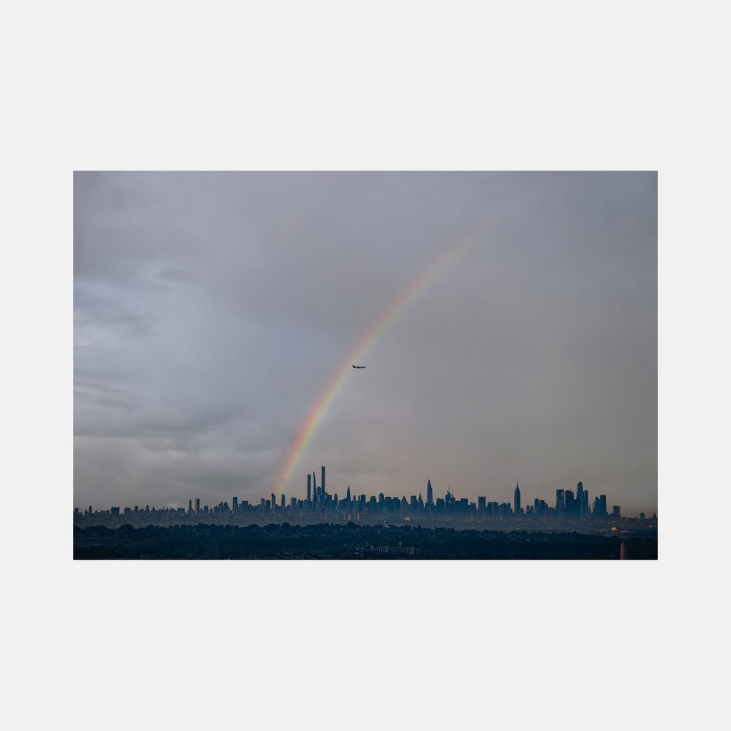 The artwork 'Chasing Rainbows' by Dennis Maida captures a dramatic urban landscape featuring a vibrant rainbow arching over the Manhattan skyline. The photograph showcases moody gray storm clouds contrasting with the city's silhouetted skyscrapers against a dusky sky. A commercial airplane intersects the rainbow, adding scale and movement to this atmospheric cityscape photograph.