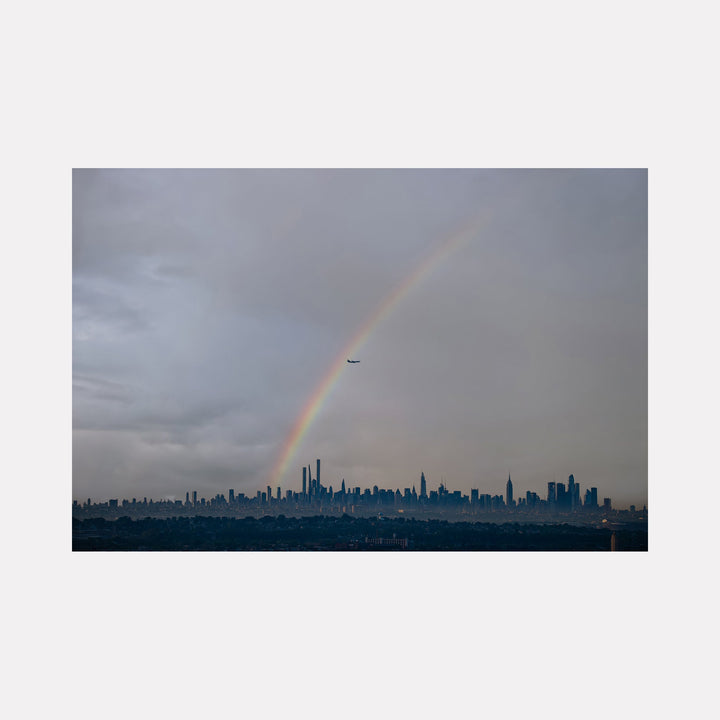 The artwork 'Chasing Rainbows' by Dennis Maida captures a dramatic urban landscape featuring a vibrant rainbow arching over the Manhattan skyline. The photograph showcases moody gray storm clouds contrasting with the city's silhouetted skyscrapers against a dusky sky. A commercial airplane intersects the rainbow, adding scale and movement to this atmospheric cityscape photograph.