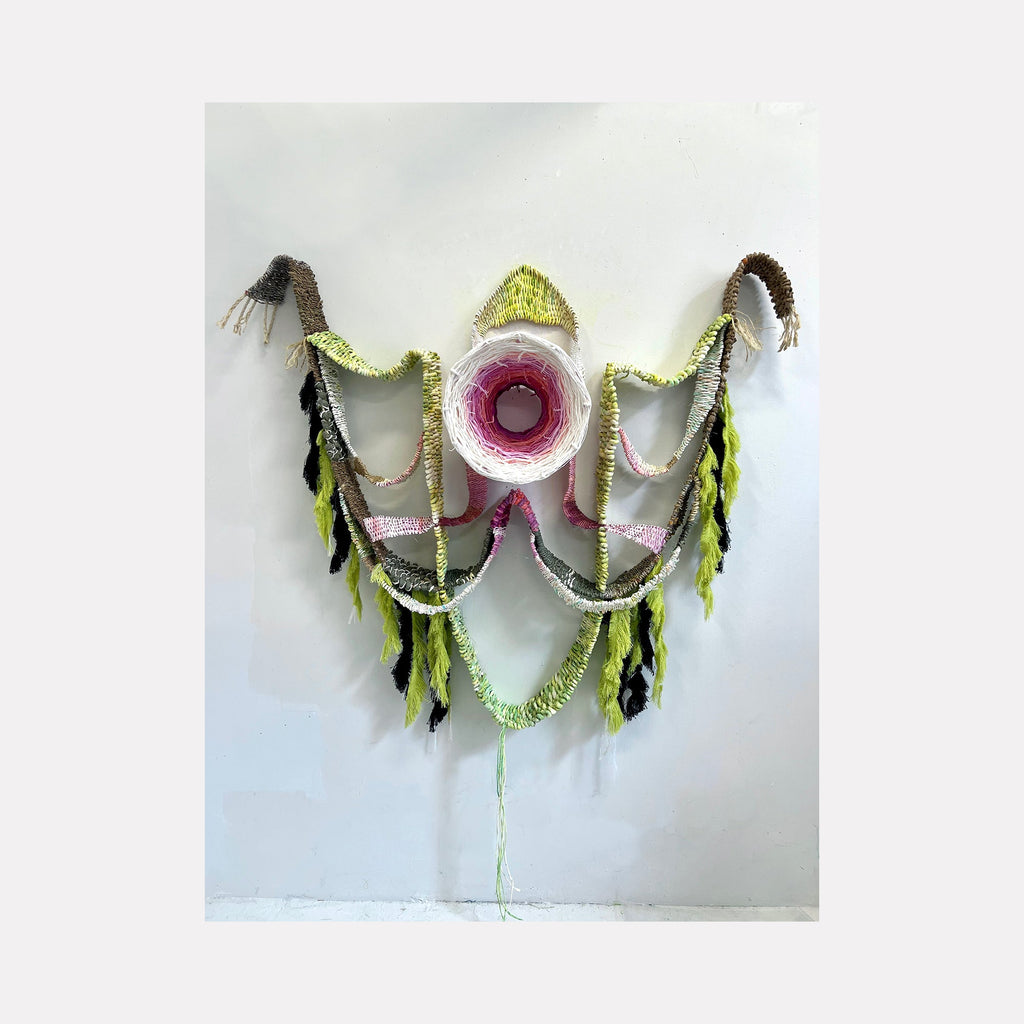 The artwork 'Emergence' by Kathie Halfin features a symmetrical fiber wall sculpture with a central pink circular form radiating outward through draped textile elements. Crafted from sisal, cotton, and hand-dyed paper yarn, the piece combines lime green and dark brown accents with flowing garland-like strands creating an organic, wing-like formation against a white background by Kathie Halfin.