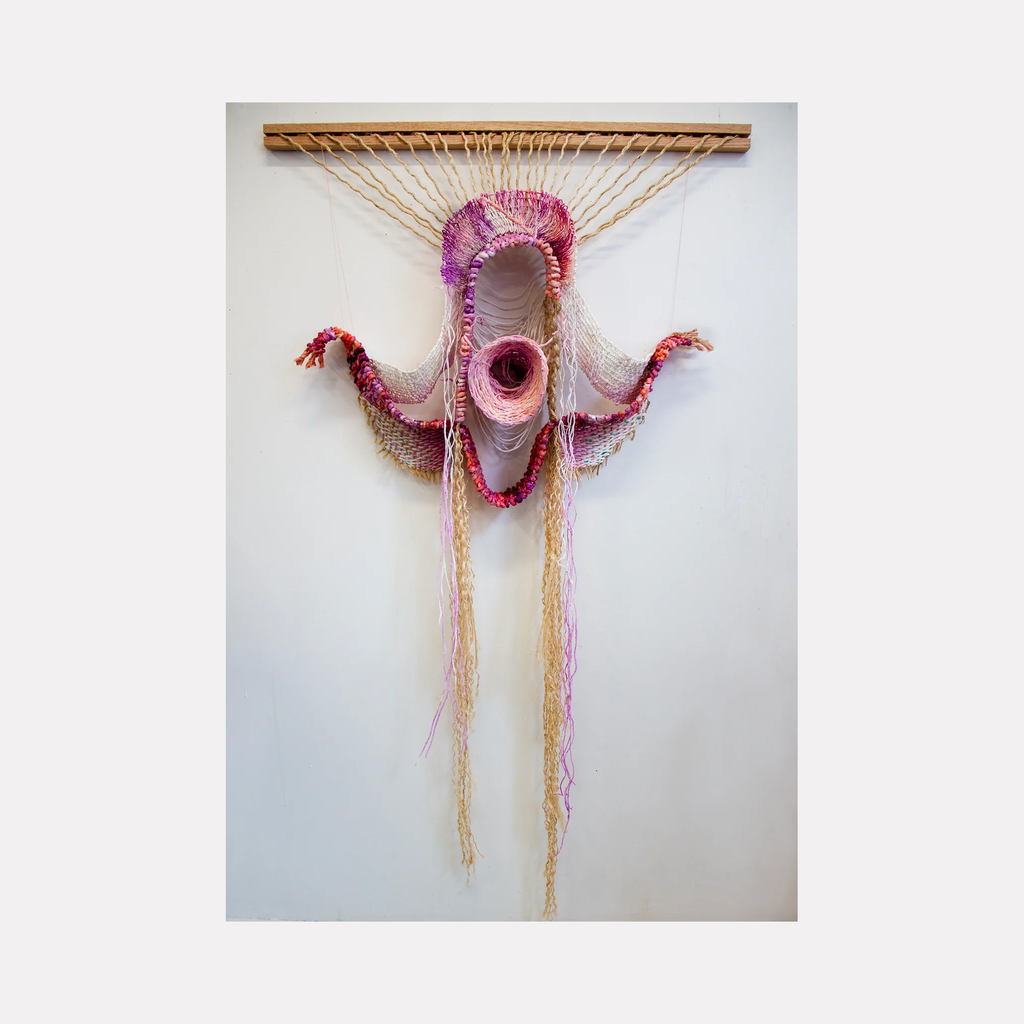 The artwork 'The Fire Pink Blooms Wild' by Kathie Halfin features an abstract fiber sculpture with a wing-like form in vibrant pink and beige hues. Hand-dyed paper yarn and sisal rope create organic flowing shapes suspended from a wooden beam, with a central circular opening surrounded by textured, undulating patterns that cascade downward, measuring 50x42 inches.