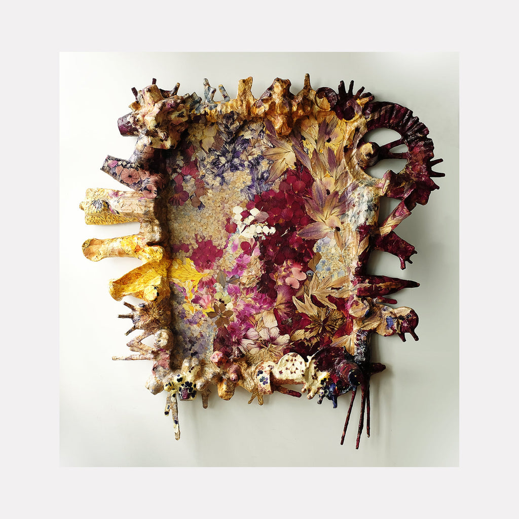 The artwork 'Framed Utopia' by Valerie Hallier features a textural mixed-media composition of preserved flower petals arranged in an abstract pattern. Deep burgundy, golden yellow, and pale pink petals create a dynamic, three-dimensional effect against a neutral canvas. Recycled packaging materials add depth and structure, while polyurethane varnish provides a protective, glossy finish.