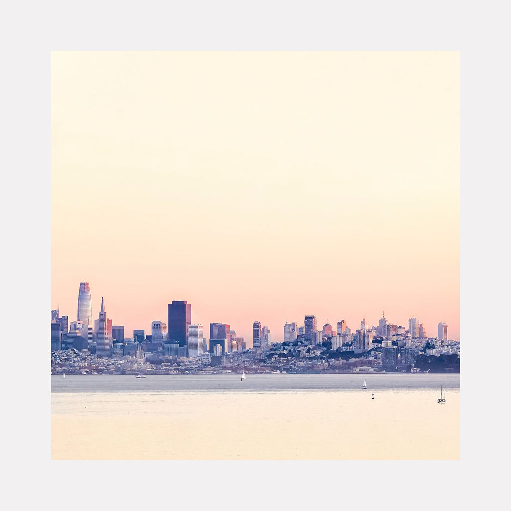 The artwork 'Golden Hour on San Francisco' by Fabienne Sowa-Dobkowski captures the city skyline at sunset, with towering skyscrapers silhouetted against a pastel pink and cream sky. The iconic downtown buildings reflect the warm light while small boats dot the tranquil bay waters below, creating a serene urban landscape photographed on archival luster paper.