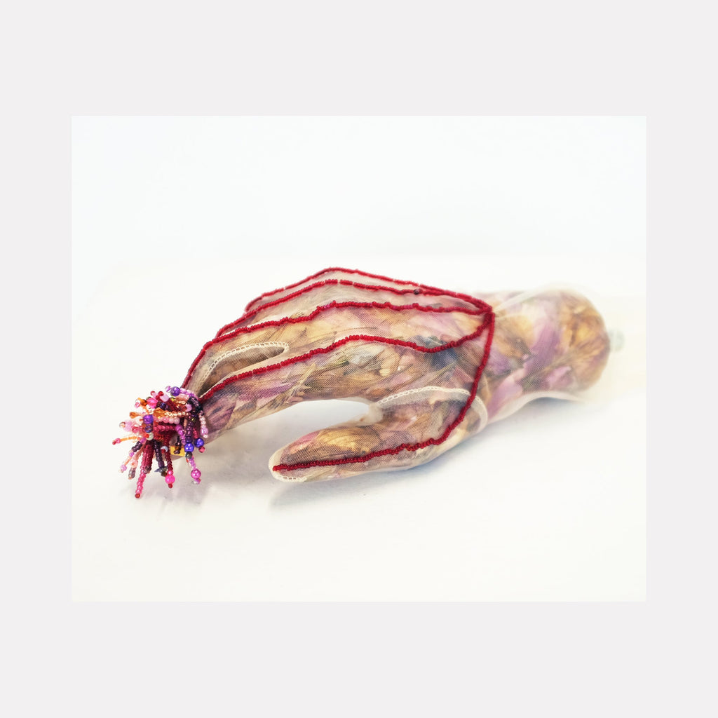 The artwork 'Indexed' by Valerie Hallier features a sculptural composition of a pale mannequin hand wrapped in delicate pantyhose, adorned with dried flower petals and vibrant red linear details. Purple and pink glass beads cluster at one end, creating a crystalline texture, while threads interweave throughout the 3.5 x 10 inch mixed media piece.