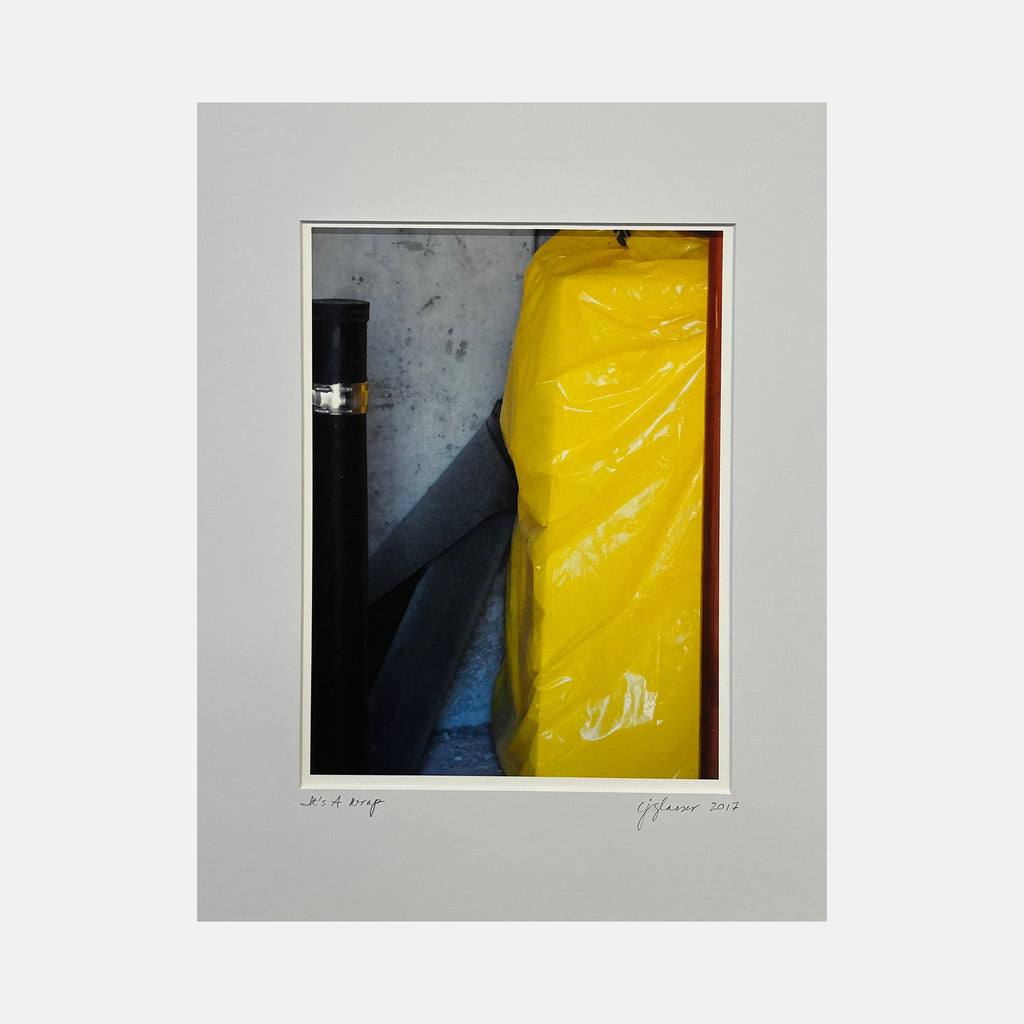 The artwork 'It's A Wrap' by Cora Jane Glasser features a striking contrast between a vibrant yellow plastic wrapping material and dark industrial elements, captured in digital photography. The composition juxtaposes glossy textures with matte surfaces, showcasing dramatic shadows and highlights on archival velvet paper. - Cora Jane Glasser